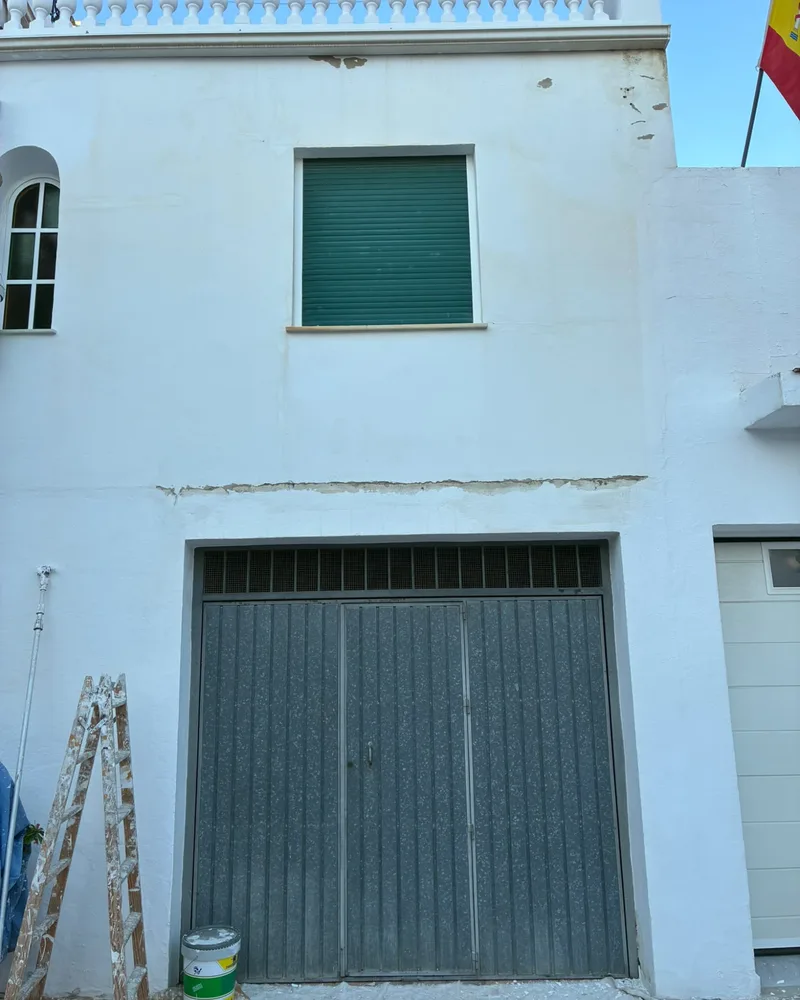 Fachada de una casa con garaje antes de pintarla, con la pintura saltada bajo la cornisa y una franja abierta sobre la puerta del garaje