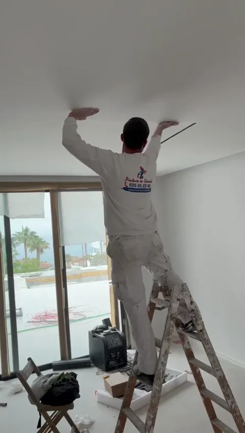 Pintor de Pinturas hermanos Carvajal subido a una escalera pintando el techo de un salón con ventanal y vistas al mar